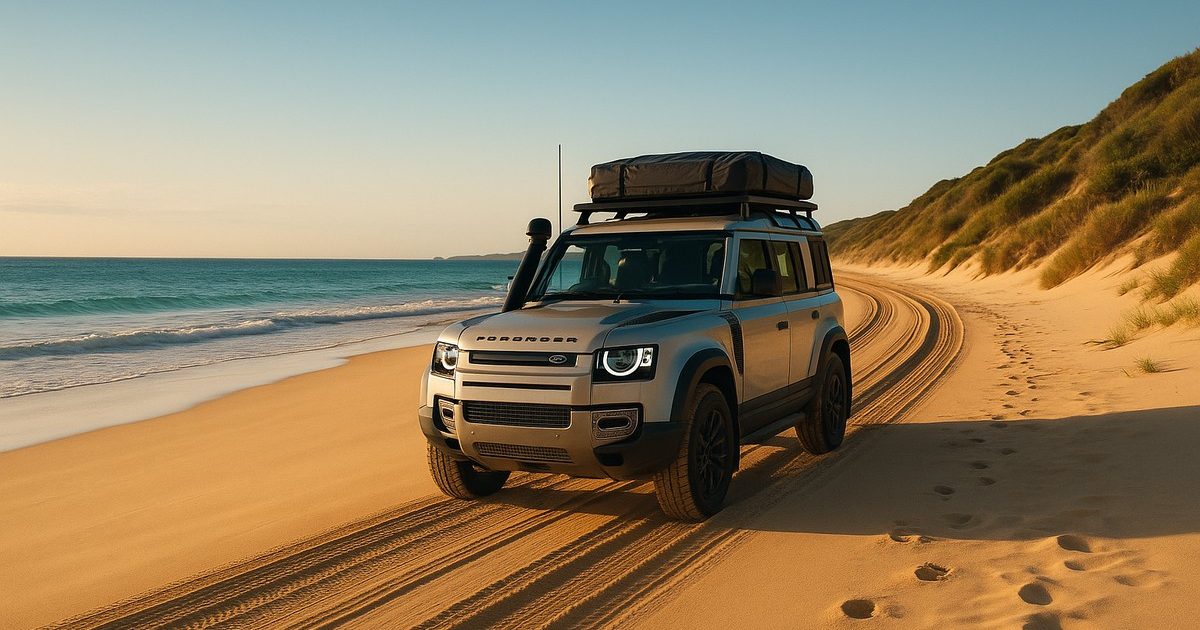Expedition-equipped Land Rover Defender driving along a secluded Australian beach at golden hour, roof rack loaded, ocean surf and sand dunes in the background